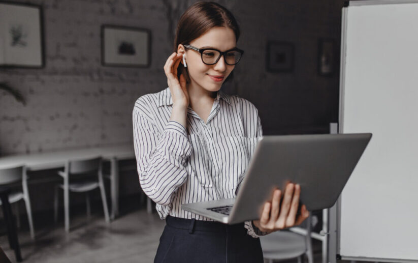 Cute girl in stylish glasses smiles, puts on wireless earphone and holds open laptop on background of office board.